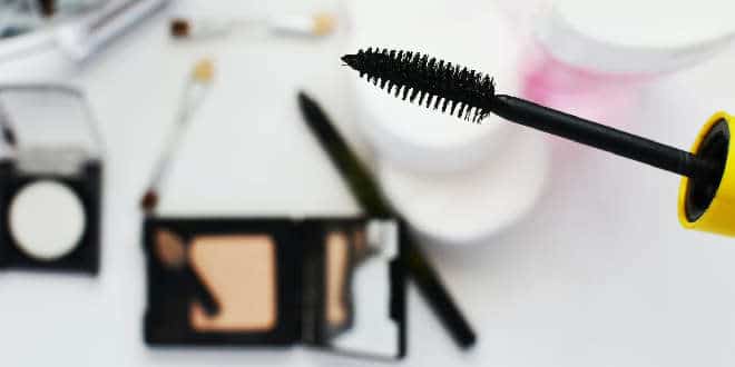 A person is holding a mascara brush in front of cosmetics.