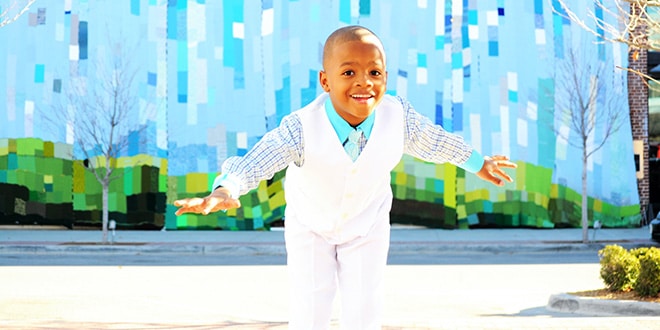 A young boy in a white vest and tie skateboarding in front of a colorful mural.